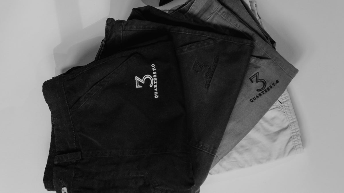 Three pairs of folded shorts stacked on a light gray background, shown in black, dark gray, and light gray. The black pair features a white embroidered logo and text reading 'QUARTERSTUDY'.
