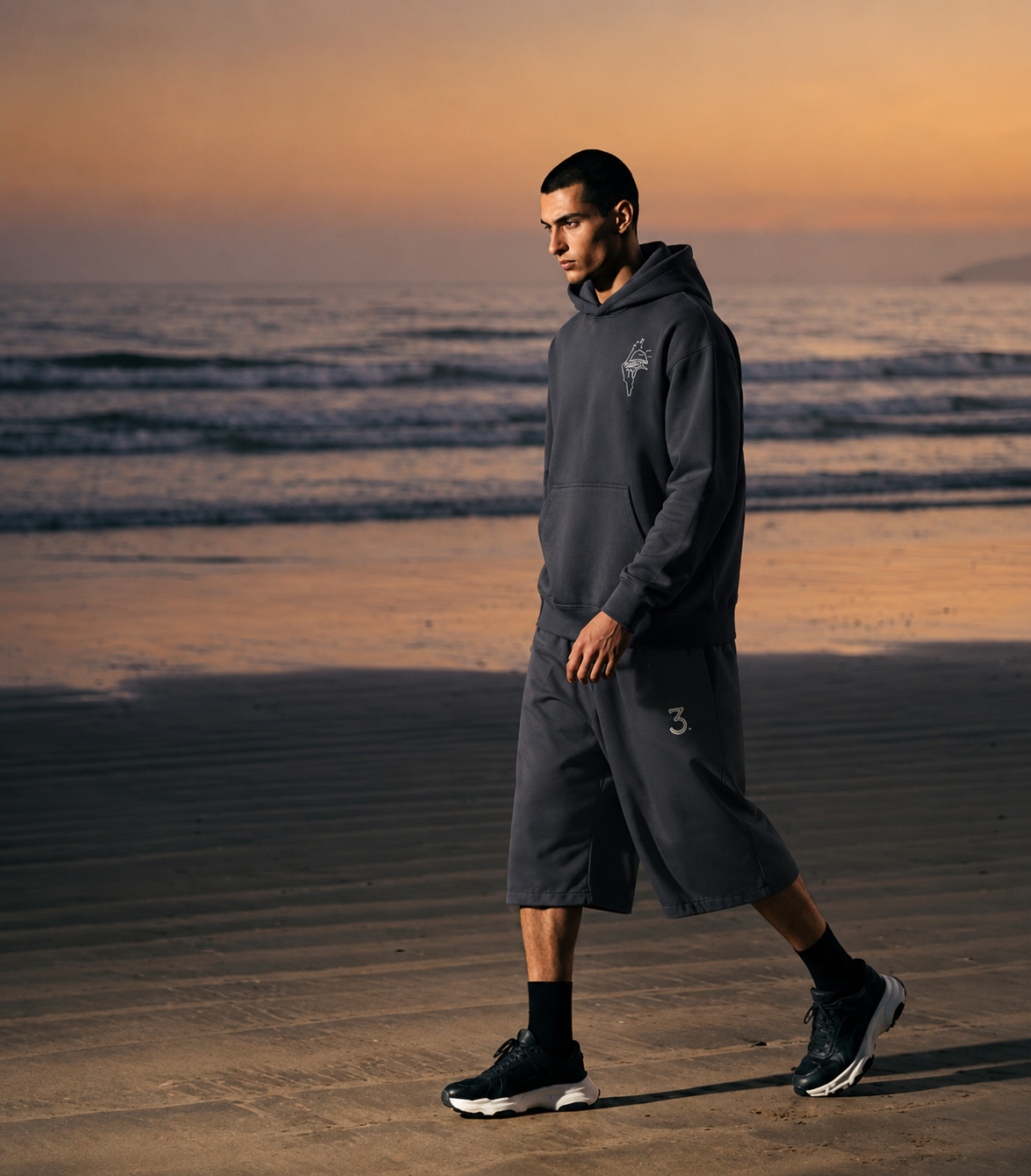 A man walks along a beach at sunset wearing a dark gray hooded sweatshirt and matching shorts, with ocean waves and a warm orange sky in the background.