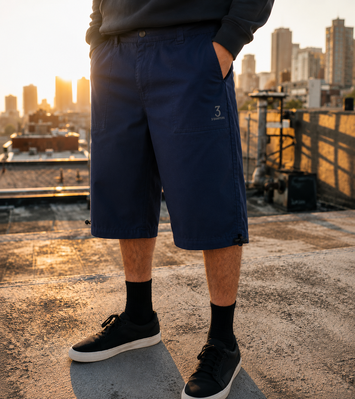 A person standing on a rooftop at sunset wearing navy knee-length shorts, a dark sweatshirt, black socks, and black sneakers with white soles. The city skyline is visible in the background.