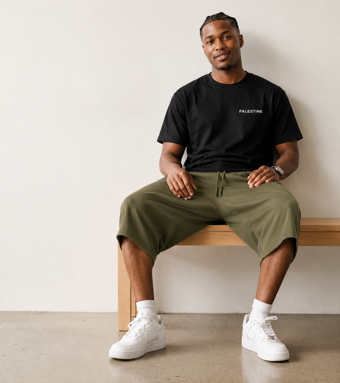 A man sits on a wooden bench wearing a black t-shirt with 'PALESTINE' printed in white on the chest, olive green drawstring shorts, and white sneakers against a plain off-white wall.